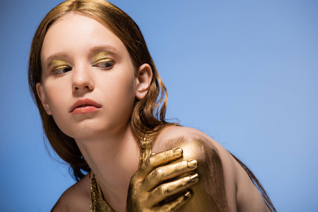 Jonge blonde model met gouden verf op de hand aanraken schouder geïsoleerd op blauw  - Foto, afbeelding