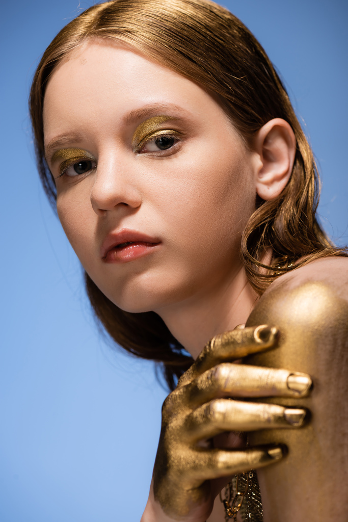Portrét mladé ženy se zlatým make-upem a malovat na ruce při pohledu na kameru izolované na modré  - Fotografie, Obrázek