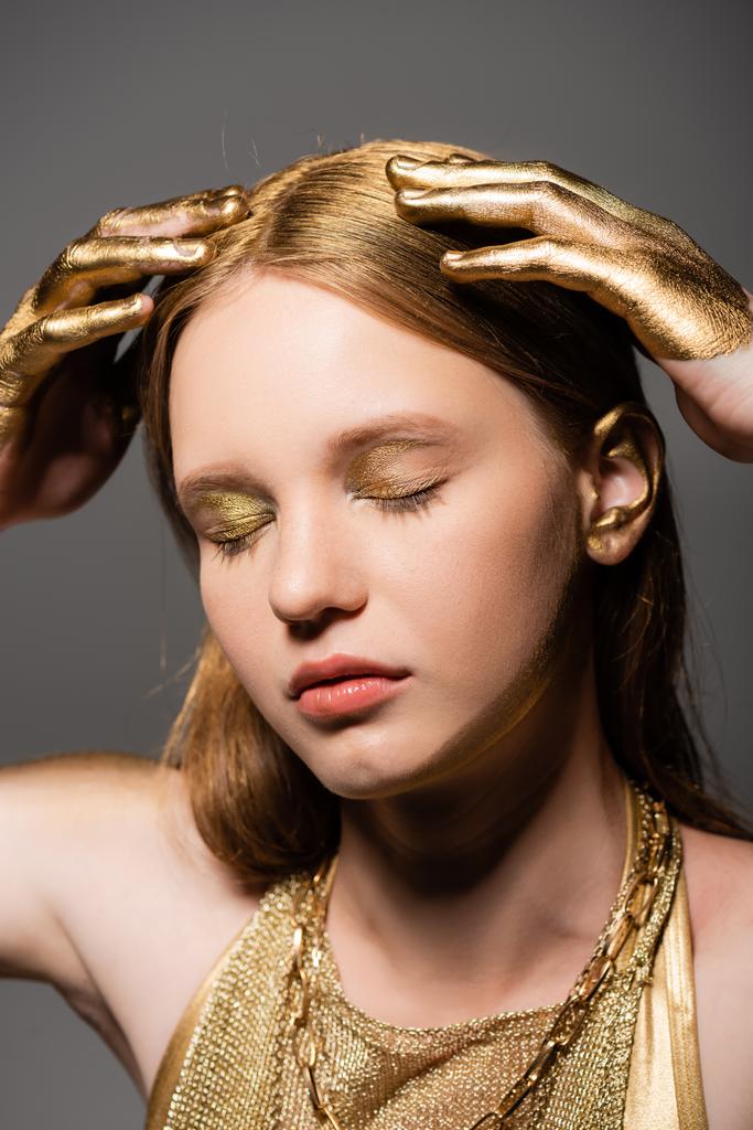 Młody model z makijażem i dłońmi w złotej farbie dotykając włosów odizolowanych na szarości  - Zdjęcie, obraz