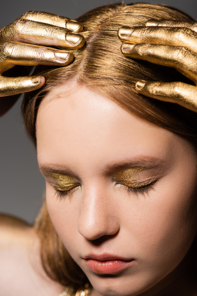 Portrait of young woman with golden makeup and paint on hands touching hair and closing eyes isolated on grey  - Photo, Image