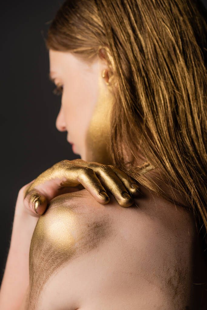 Πλευρική άποψη του burred γυναίκα με χρυσή μπογιά στο χέρι αγγίζοντας τον ώμο απομονώνονται σε γκρι  - Φωτογραφία, εικόνα