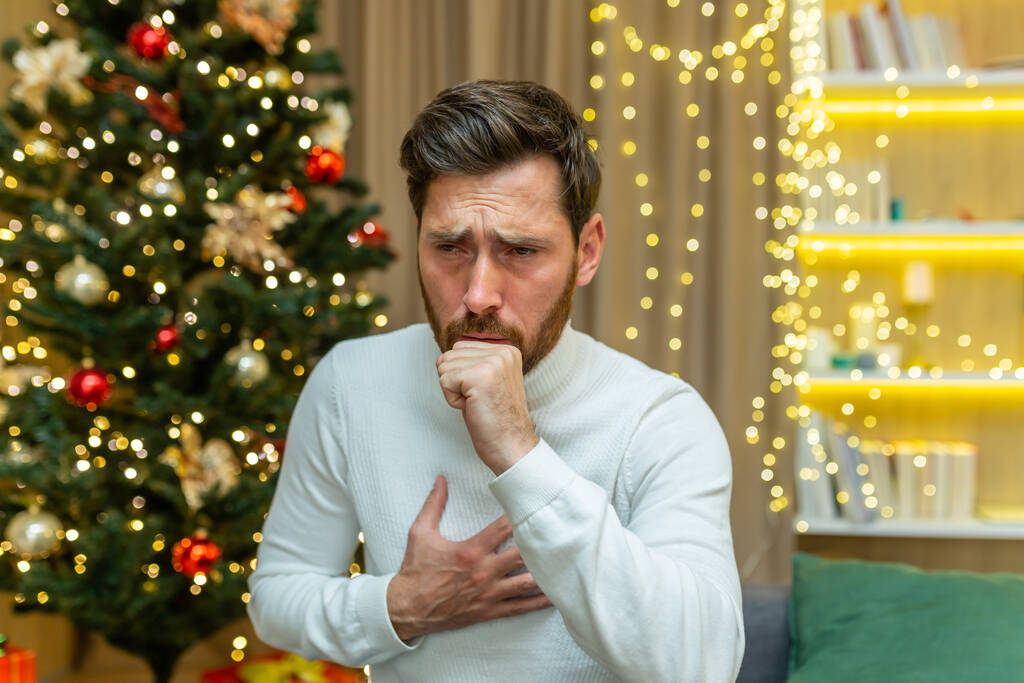 άνθρωπος βήχει τα Χριστούγεννα στο σπίτι κάθεται μόνος στον καναπέ άρρωστος τις διακοπές του νέου έτους στο σαλόνι. - Φωτογραφία, εικόνα