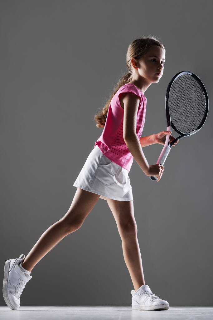 Young female tennis player in pink outfit. Little girl posing with racket and ball. Half side lit silhouette against gray background. - Photo, Image