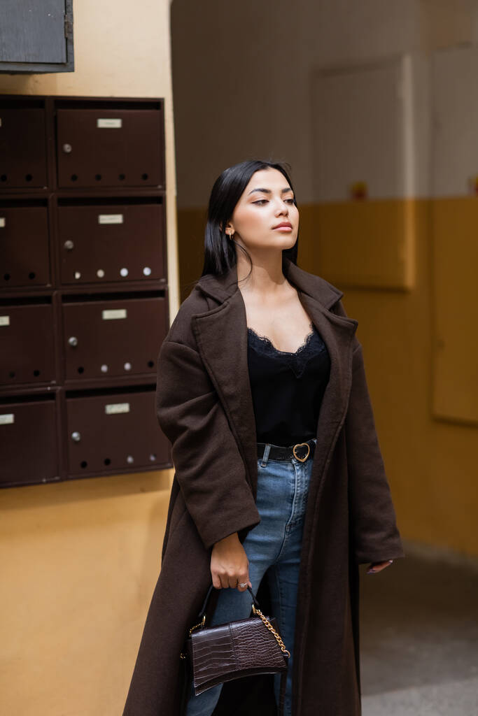 brunetta donna in cappotto alla moda in piedi con borsetta vicino edificio con cassette postali a Praga - Foto, immagini