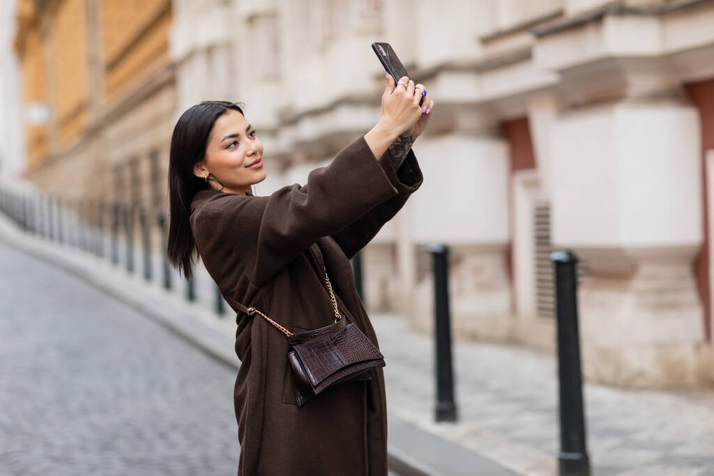 χαμογελαστή μελαχρινή γυναίκα σε μοντέρνο παλτό λαμβάνοντας selfie στο κινητό τηλέφωνο σε prague - Φωτογραφία, εικόνα
