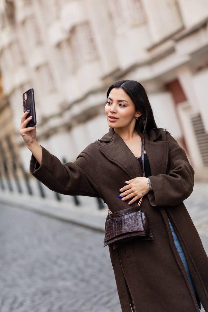 joven morena en abrigo marrón tomando selfie en calle borrosa en praga - Foto, imagen