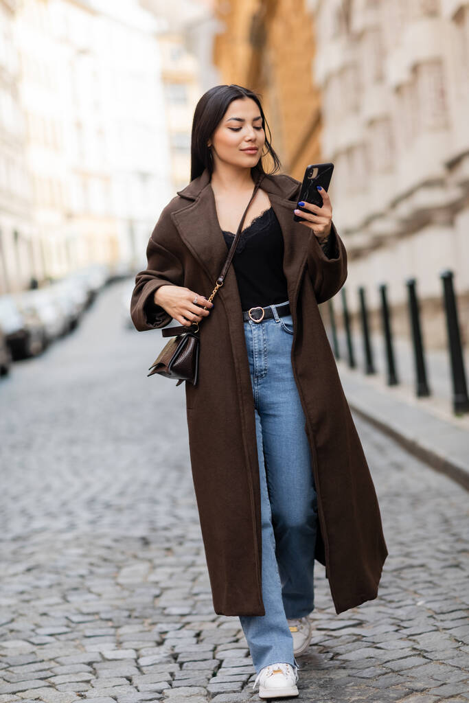 コートを着たブルネットの女性とジーンズはプラハの通りを歩いている間にスマートフォンを見て - 写真・画像