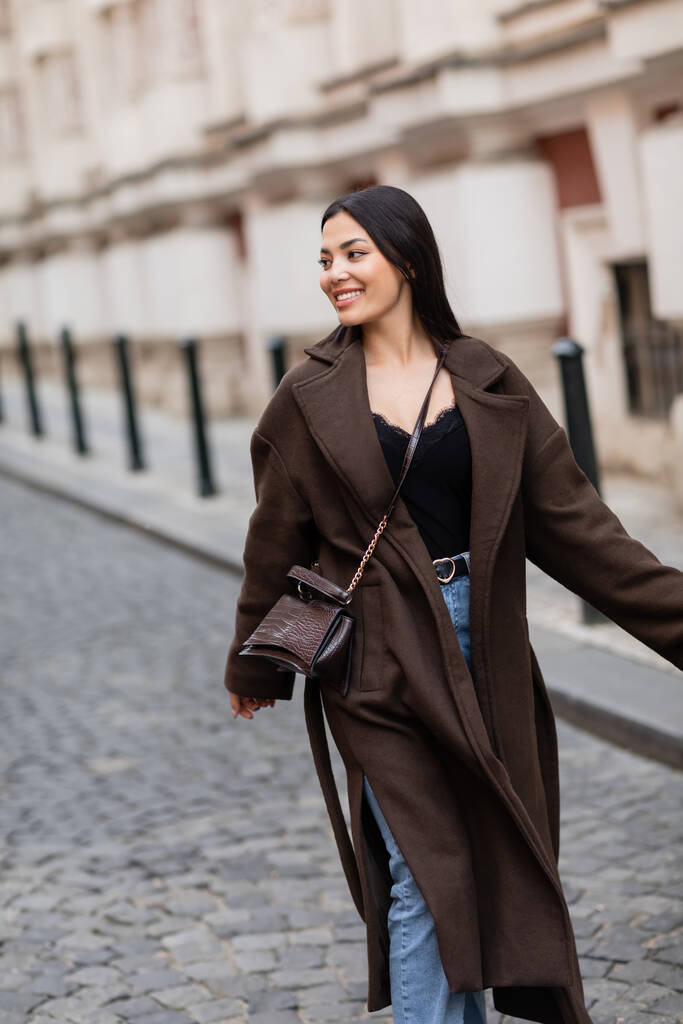 wesoła brunetka w stylowym płaszczu z krzyżówką spacerująca po ulicy miejskiej w Pradze - Zdjęcie, obraz