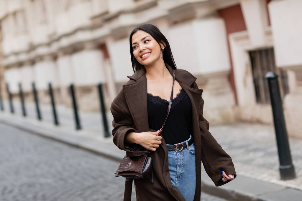 glimlachende brunette vrouw in trendy jas met crossbody lopen op straat in Praag - Foto, afbeelding