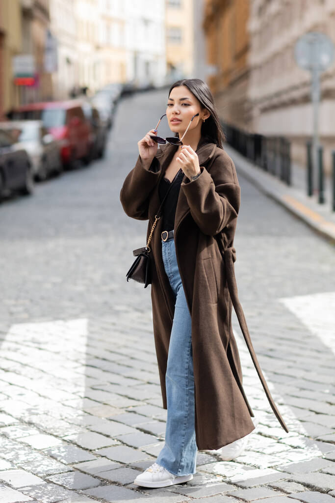 pleine longueur de jolie femme brune en manteau tenant des lunettes de soleil sur la rue de la ville dans la prague - Photo, image