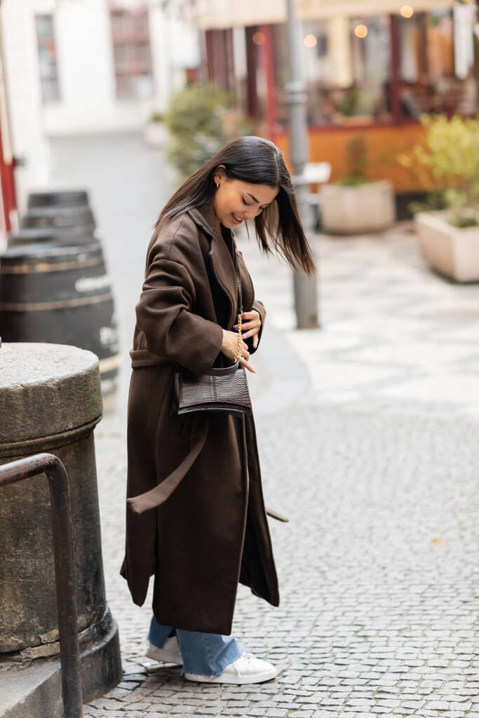 piena lunghezza di donna sorridente in marrone cappotto apertura crossbody mentre in piedi su strada a Praga - Foto, immagini