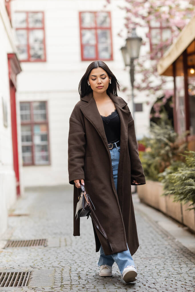 volledige lengte van stijlvolle brunette vrouw in draadloze oortelefoons lopen op smalle straat in Praag - Foto, afbeelding
