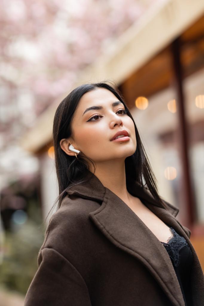 young brunette woman in brown coat looking away while listening music in wireless earphone on street in prague - Photo, Image