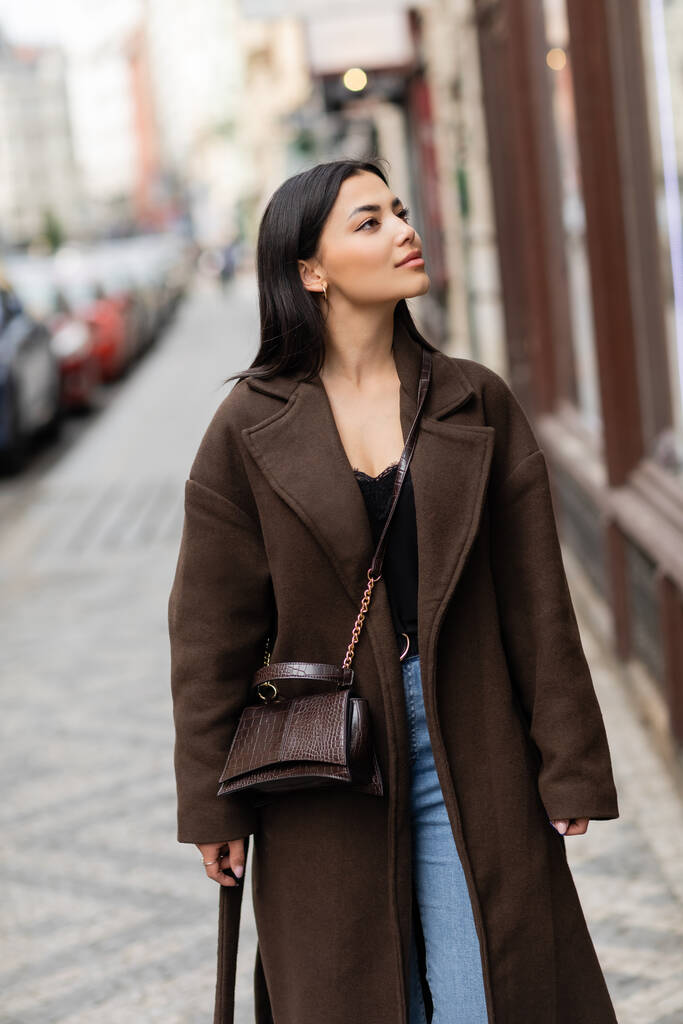 かなりブルネット女性で茶色いコートでトレンディーなクロスボディと離れてプラハの街を見て - 写真・画像