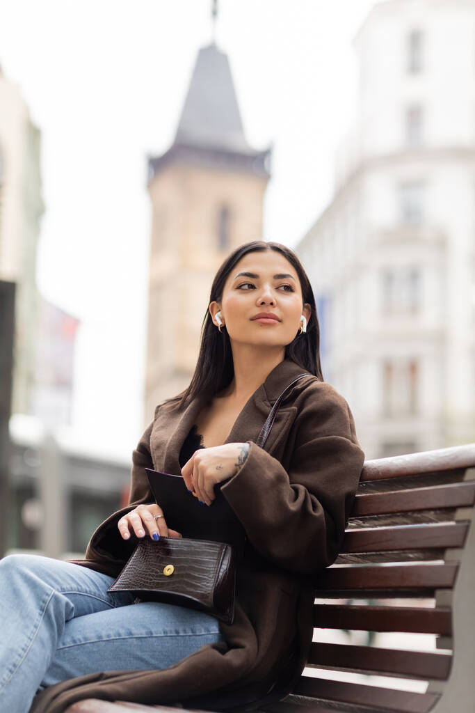 mujer joven en auriculares inalámbricos abriendo bolso de moda y mirando hacia otro lado en el banco en prague - Foto, imagen