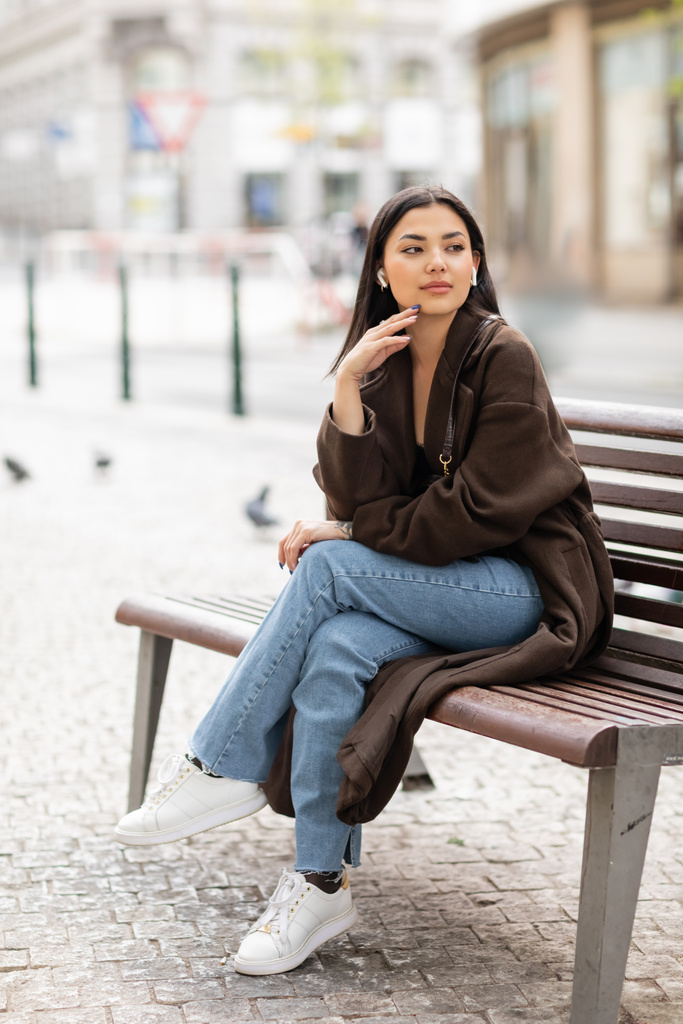 pretty woman in trendy autumn clothes listening music in wireless earphones on bench  in prague and looking away - Photo, Image