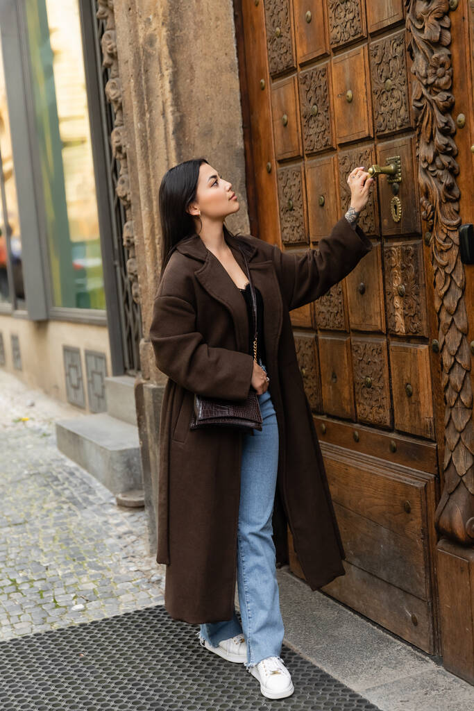 comprimento total da mulher morena em casaco elegante com abertura crossbody esculpida porta de madeira do edifício em prague - Foto, Imagem