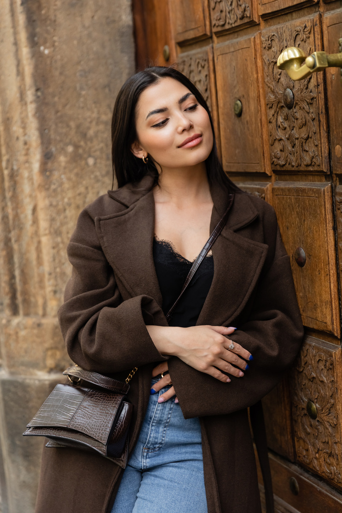 positive brunette woman in stylish autumn coat standing near carved wooden door in prague - Photo, Image