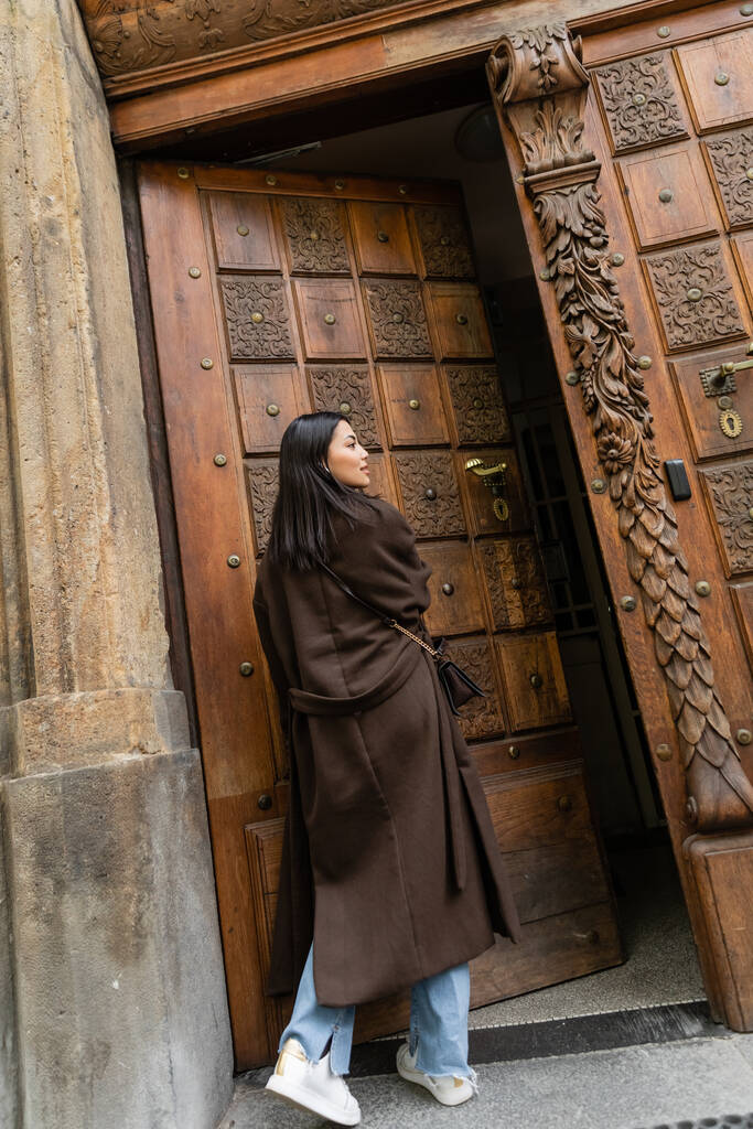 fiatal barna nő barna kabátban besétál nyitott fa ajtó Prágában - Fotó, kép