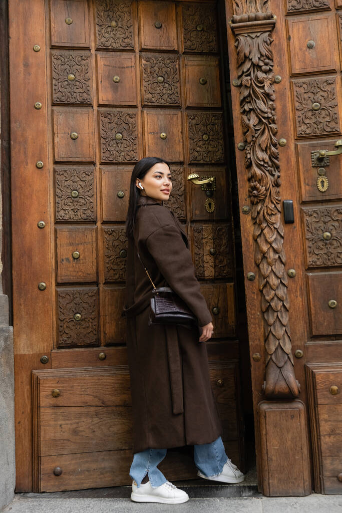 woman in coat and wireless earphone looking away near carved door in prague - Photo, Image