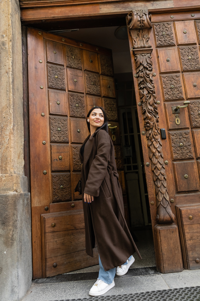 pełna długość stylowa kobieta w brązowym płaszczu uśmiechnięta i patrząc daleko w pobliżu drewnianych drzwi w Pradze - Zdjęcie, obraz