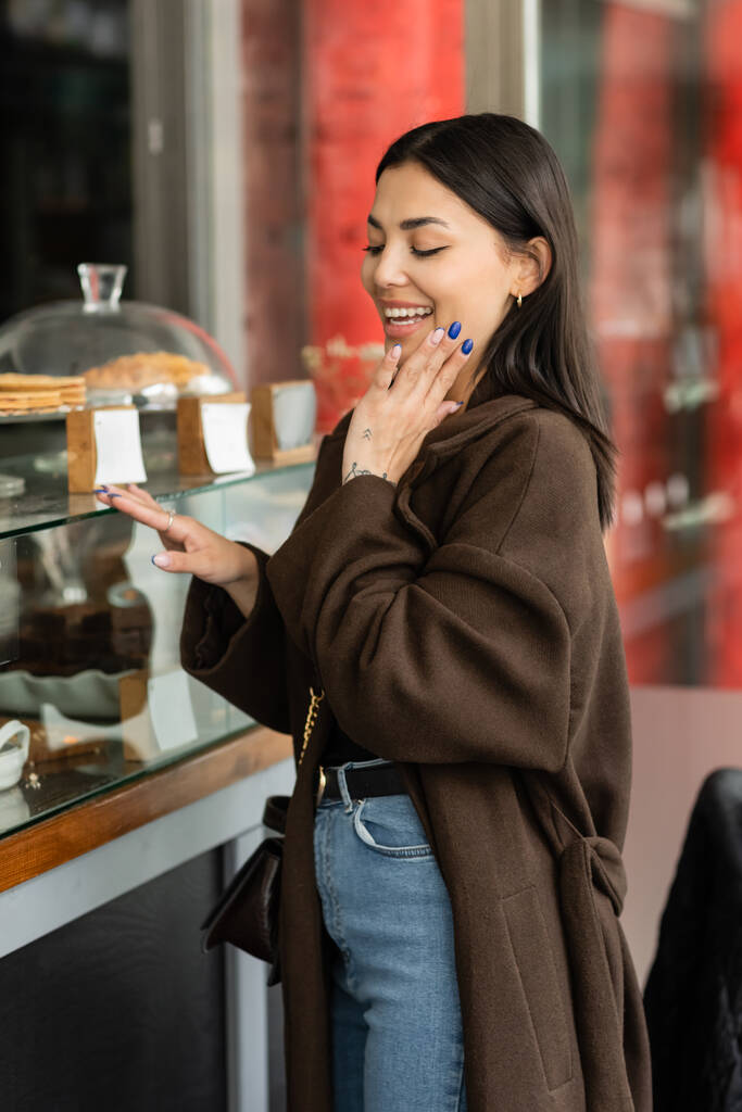 mujer asombrada sosteniendo la mano cerca de la cara mientras mira escaparate de pastelería con postres en praga  - Foto, imagen