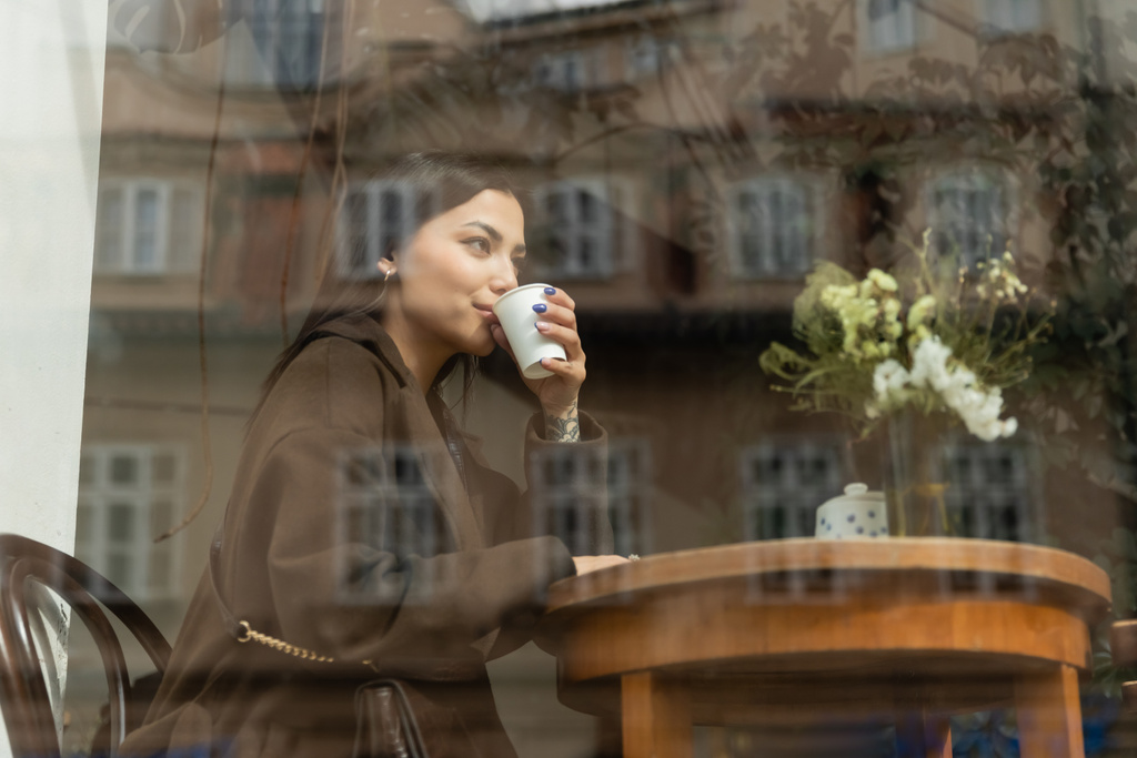 młoda kobieta w jesiennym płaszczu pije kawę w pobliżu okna w kawiarni Praga - Zdjęcie, obraz
