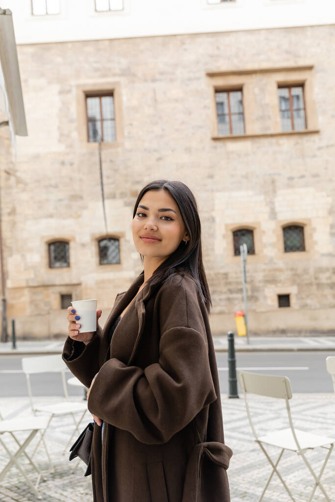 Portrait de jeune femme en manteau tenant une tasse en papier et souriant à la caméra dans la rue à Prague  - Photo, image