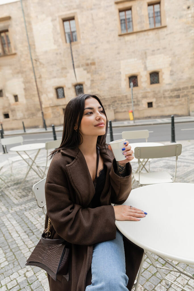 Brunette woman in coat holding paper cup with coffee in outdoor cafe on street in Prague  - Photo, Image