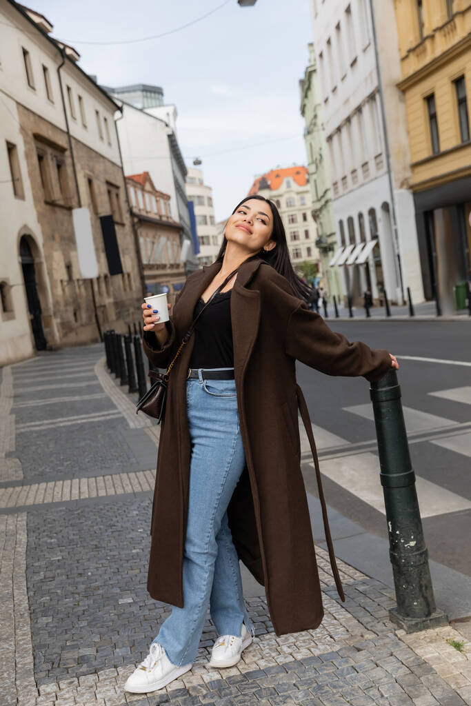 Mujer alegre en abrigo sosteniendo taza de papel en la calle urbana de Praga  - Foto, Imagen