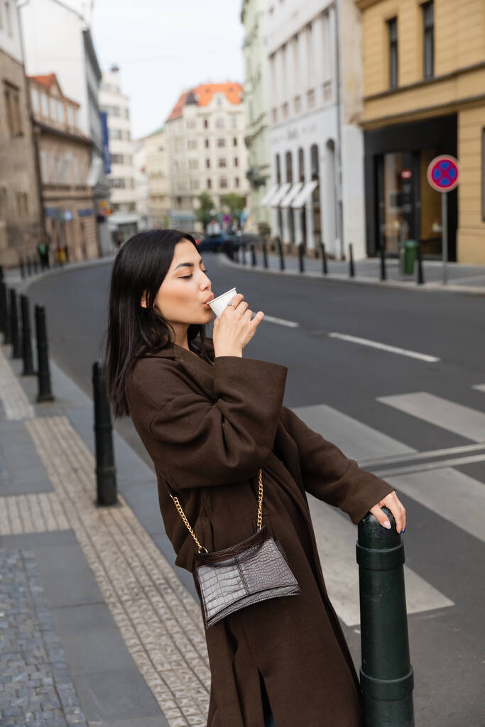 Κομψή γυναίκα σε παλτό πίνοντας καφέ από το κύπελλο χαρτί στον αστικό δρόμο στην Πράγα  - Φωτογραφία, εικόνα