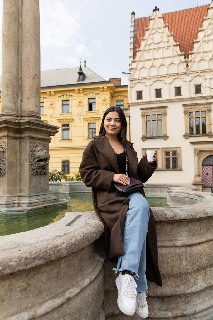 Boldog utazó kabátban tartó papír pohár közelében ősi szökőkút Károly téren Prágában  - Fotó, kép