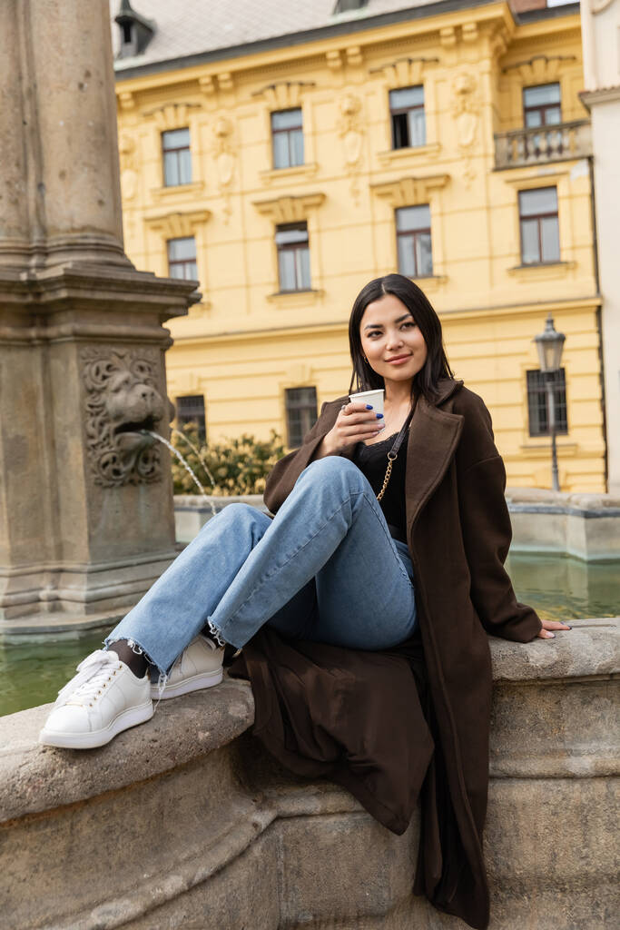 Stylová mladá žena v kabátě drží papírový kelímek u staré kašny na Karlově náměstí v Praze  - Fotografie, Obrázek