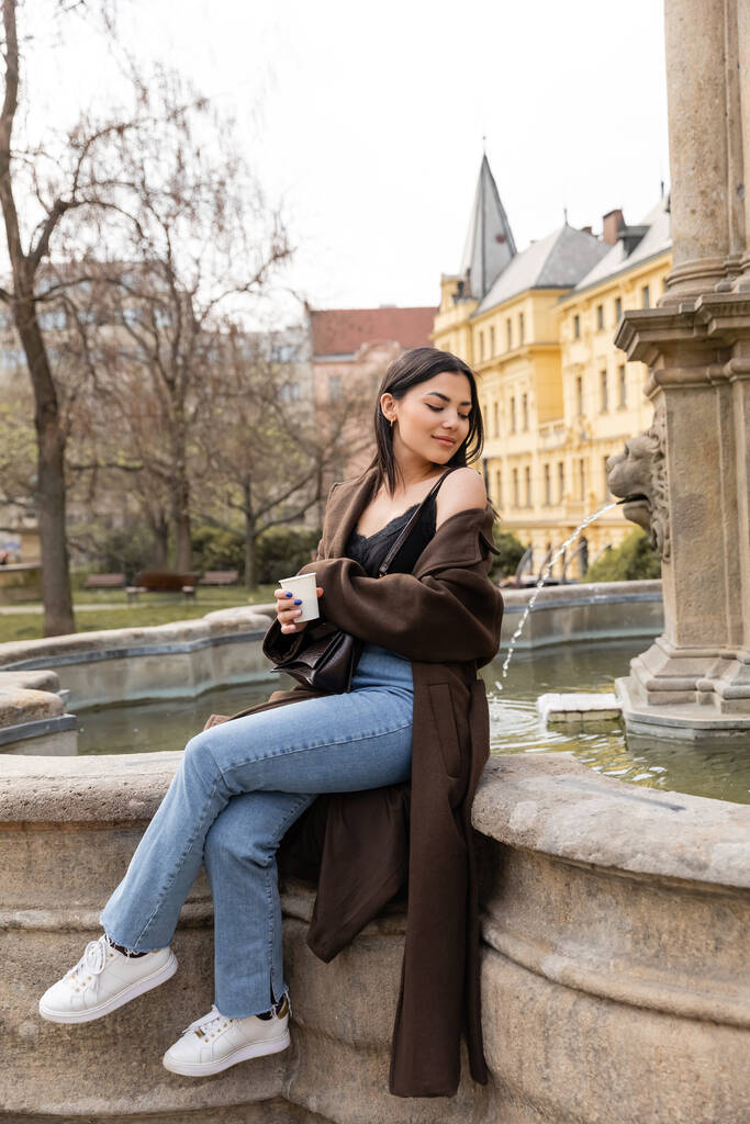 Młoda kobieta w płaszczu i dżinsach trzyma papierowy kubek przy fontannie na Placu Karola w Pradze  - Zdjęcie, obraz