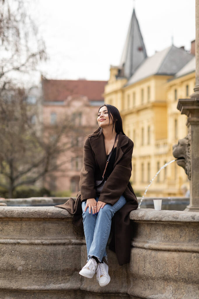 Улыбающаяся женщина в коричневом пальто смотрит вдаль возле бумажной чашки на Карловой площади в Праге  - Фото, изображение