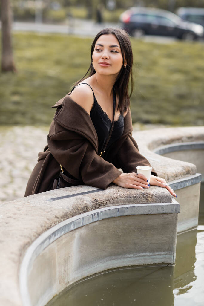 Молода жінка в коричневому пальто тримає паперову чашку біля фонтану в Празі.  - Фото, зображення