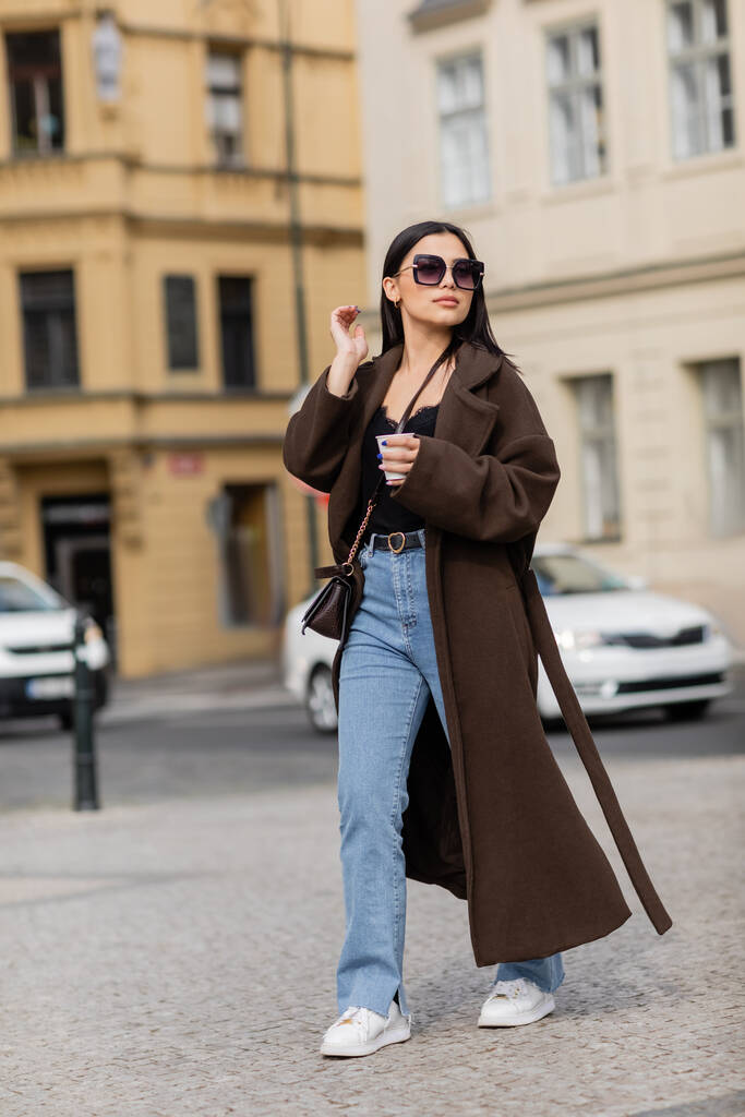 Turista de moda en abrigo y gafas de sol sosteniendo taza de papel mientras camina por la calle en Praga  - Foto, Imagen