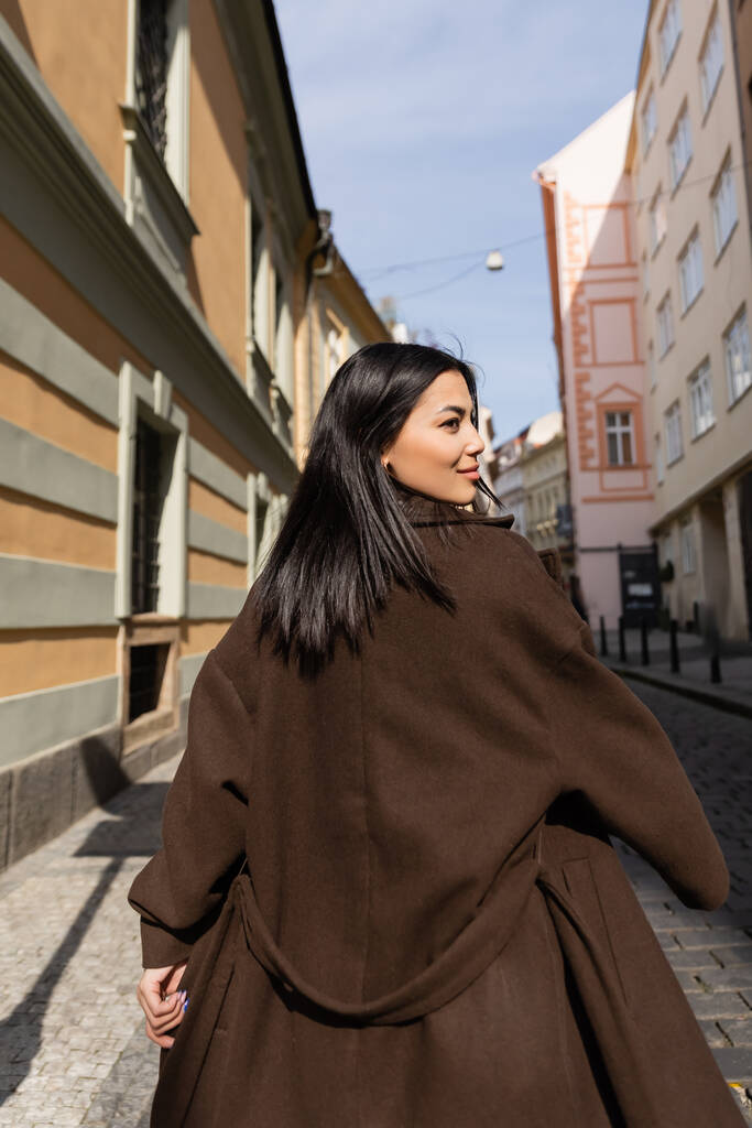 Vista lateral de mujer morena en abrigo caminando por la calle en Praga  - Foto, imagen