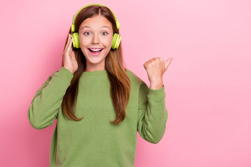 Fotografie lesklý dojem školačka nosit zelené mikiny sluchátka ukazující palec prázdný prostor izolované růžové barvy pozadí. - Fotografie, Obrázek