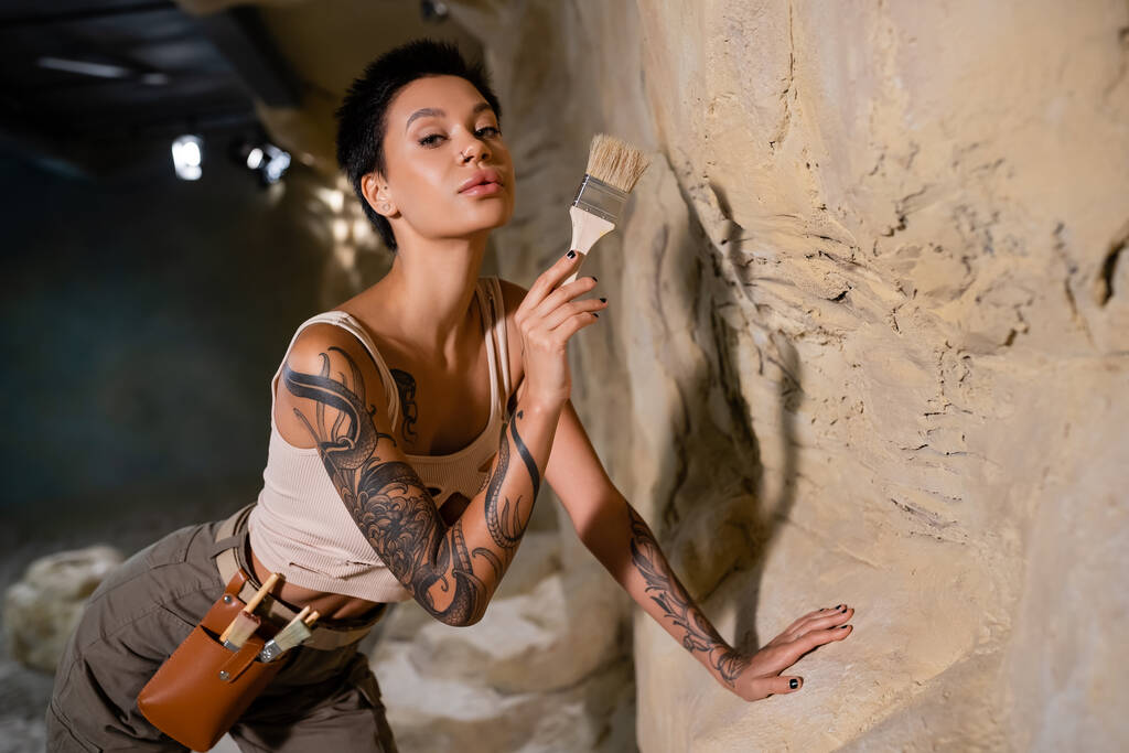 tetovaný a sexy archeolog při pohledu na kameru při držení štětce u skály v jeskyni - Fotografie, Obrázek