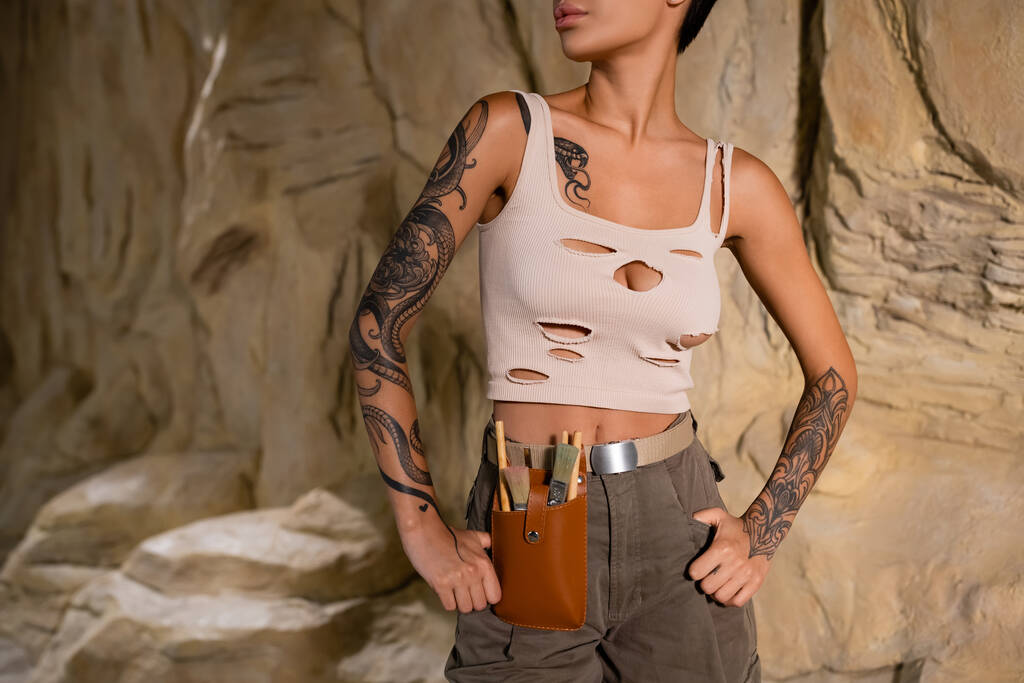 gedeeltelijke weergave van getatoeëerde archeoloog in tank top en taille tas met borstels staan met de handen op heupen in de buurt van rots - Foto, afbeelding