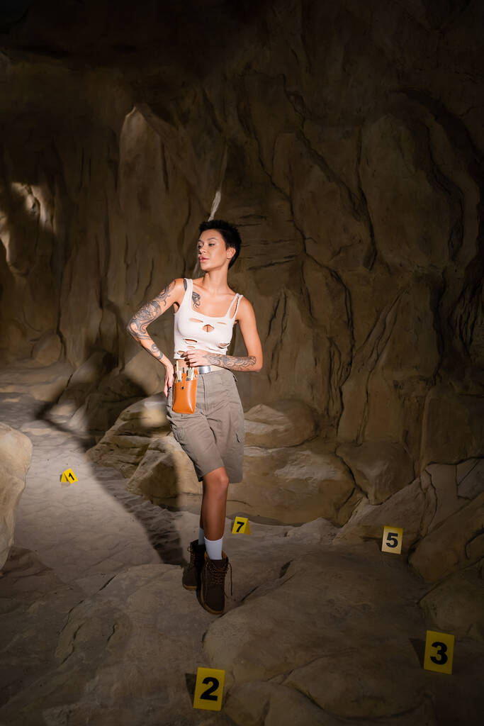 in voller Länge sexy Archäologin in Tank Top steht neben nummerierten Karten in Höhle - Foto, Bild