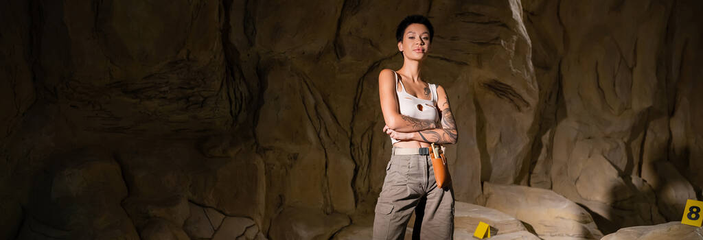 jovem arqueólogo morena em shorts e top tanque de pé com braços cruzados em caverna escura, bandeira - Foto, Imagem