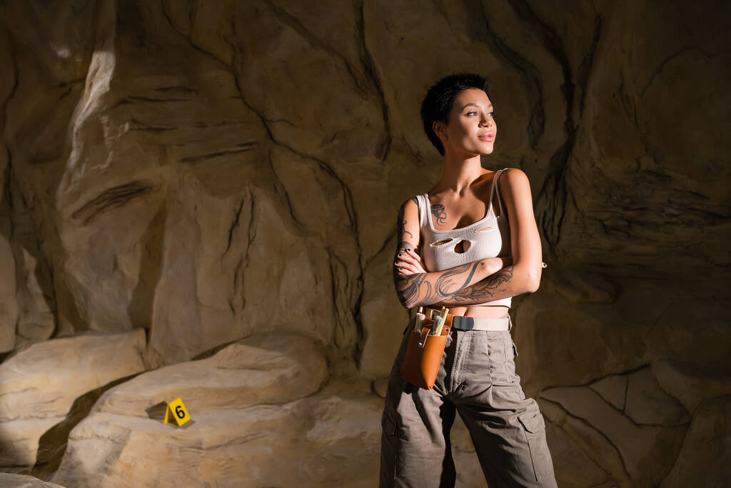 young and sexy archaeologist in tank top standing in cave with crossed arms and looking away - Photo, Image