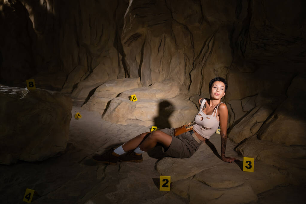 młody brunetka archeolog siedzi w pobliżu ponumerowane karty w ciemnej jaskini i odwracając wzrok - Zdjęcie, obraz