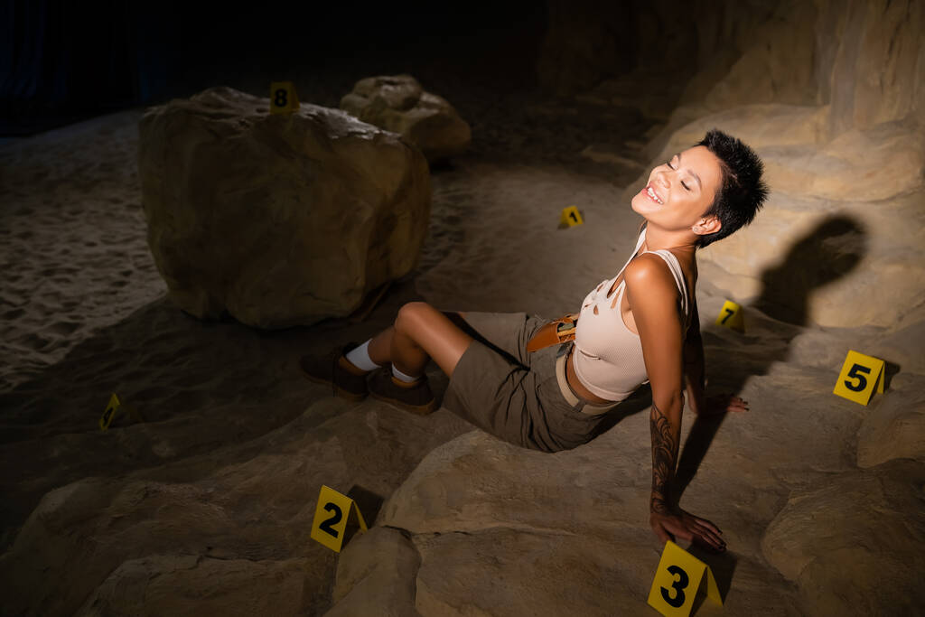 alegre arqueólogo con los ojos cerrados sentado cerca de tarjetas numeradas en la cueva oscura del desierto - Foto, imagen