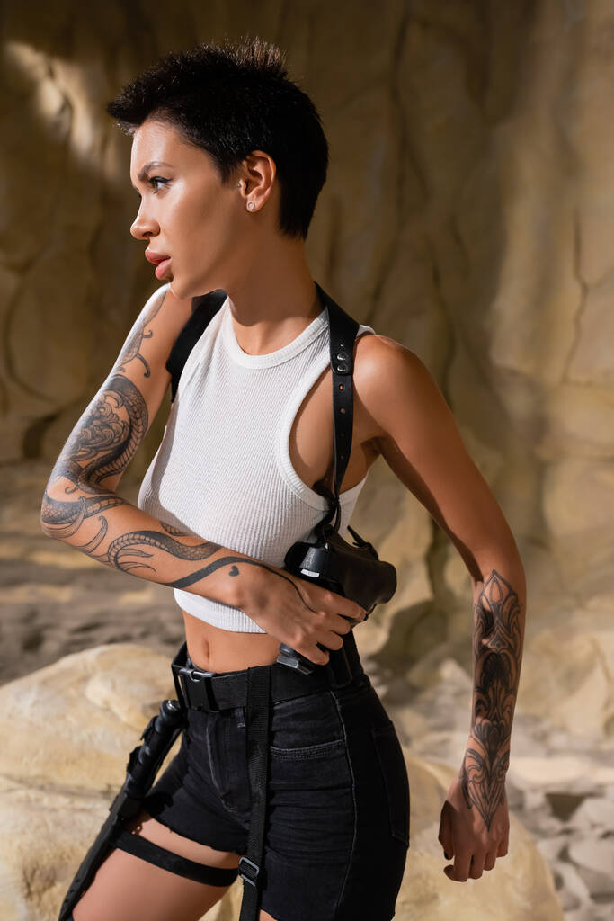 tetovaný archeolog s krátkými vlasy vytahuje zbraň z pouzdra v jeskyni - Fotografie, Obrázek