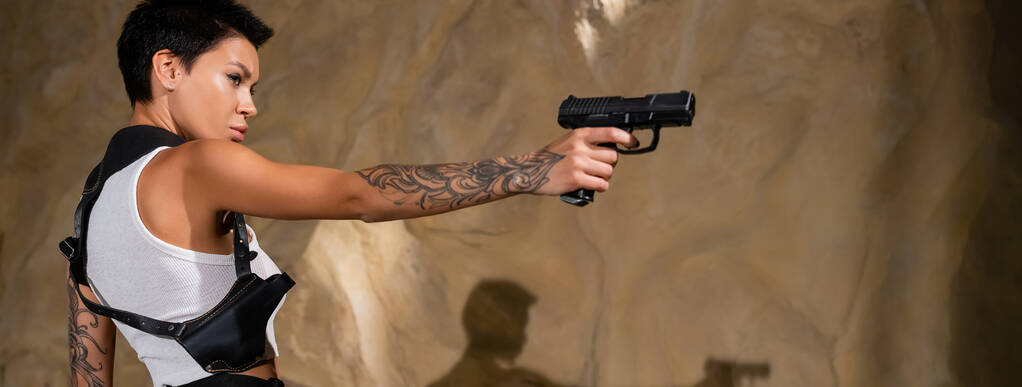 vista lateral del arqueólogo tatuado con funda sosteniendo pistola en el desierto, pancarta - Foto, Imagen