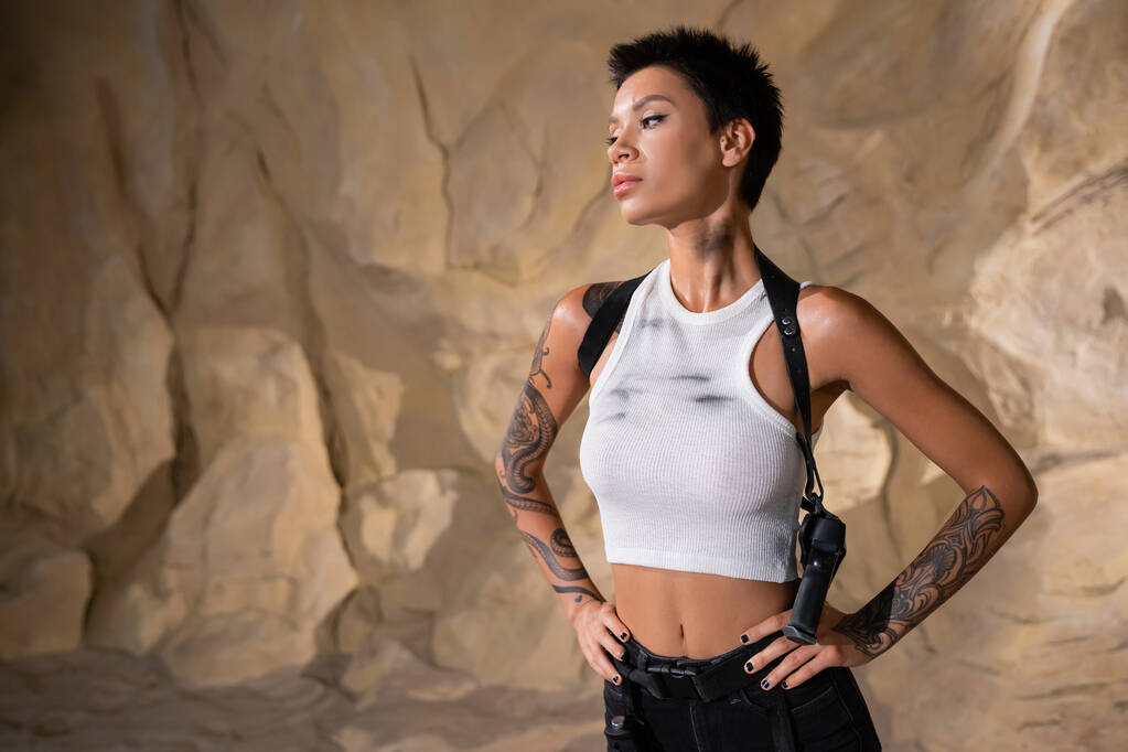 armed and tattooed archaeologist with dirty crop top standing with hands on hips - Photo, Image