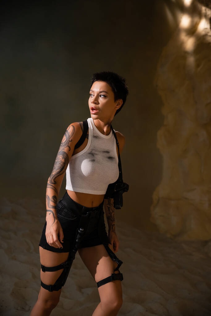 curious and tattooed archaeologist in sexy outfit with gun in holster walking in cave - Photo, Image